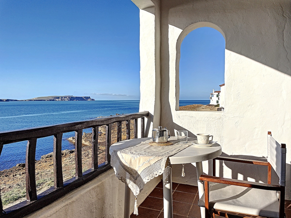 A serene balcony overlooks the sparkling sea, featuring a small table set with a teapot and cup. A single wooden chair complements the space, and a charming arched window allows natural light to fill the area, enhancing the coastal view.