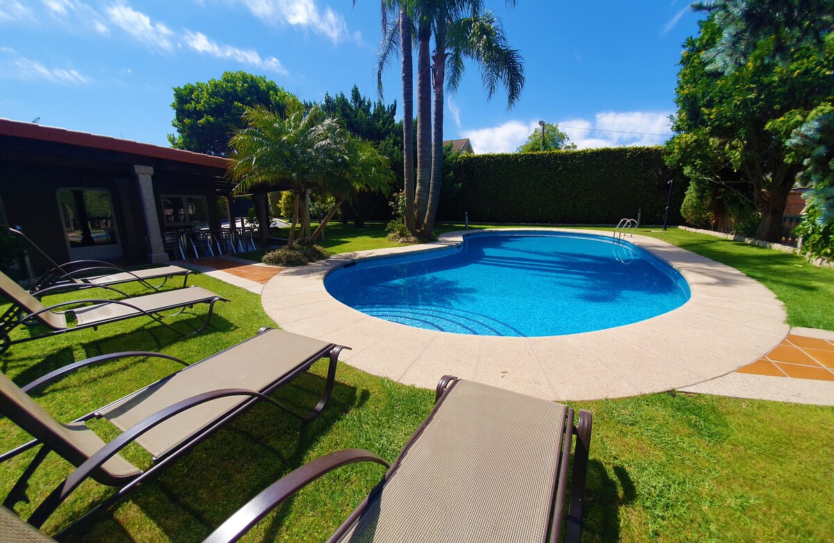 A circular pool is surrounded by well-maintained grass and palm trees. Reclining chairs are positioned along the poolside, providing ample space for relaxation. The clear blue water reflects the bright sky, and lush greenery borders the area, contributing to a peaceful outdoor setting.