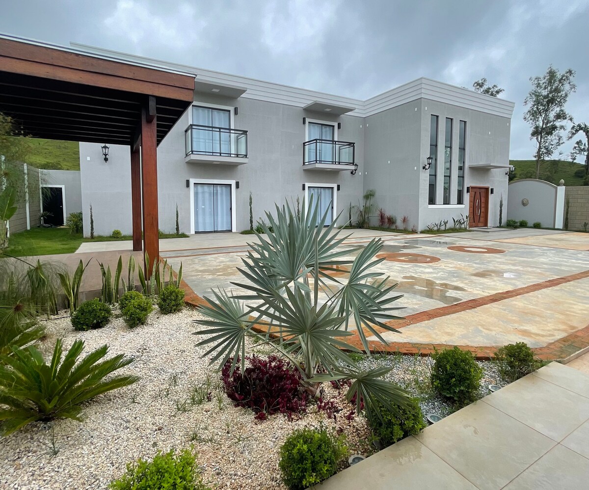 The exterior showcases a modern building with a clean facade, featuring large windows and balconies. A symmetrical stone pathway adorned with landscaping leads to the entrance. Lush greenery and ornamental plants punctuate the area, enhancing the overall aesthetic.