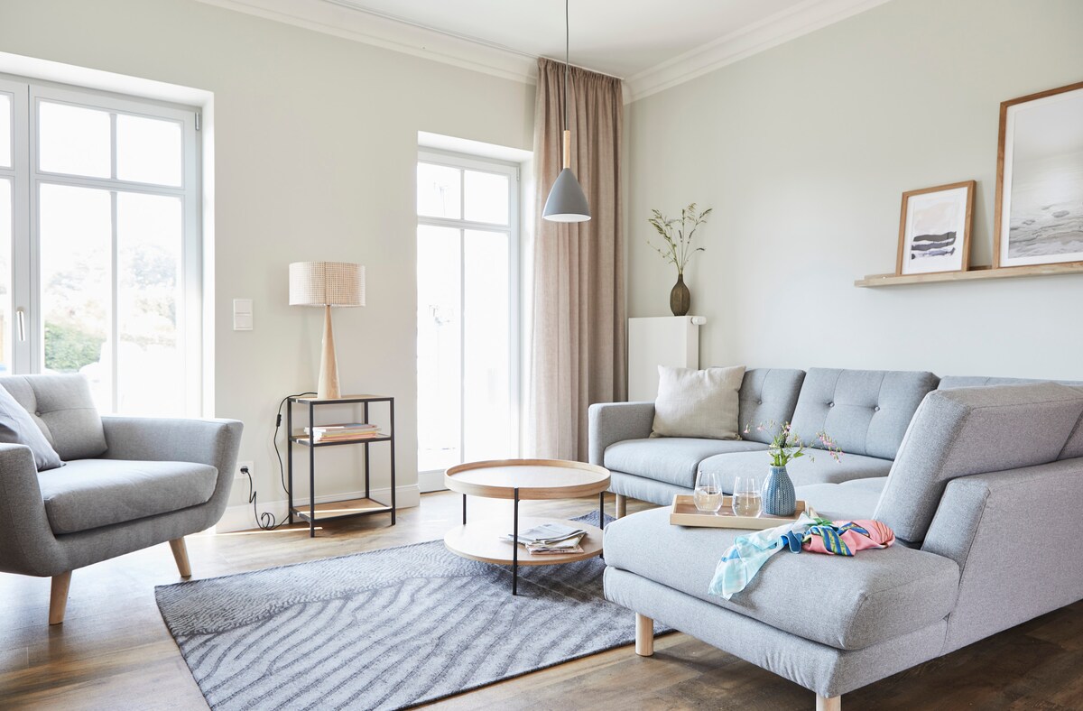 A bright living area features a comfortable gray sectional sofa and a matching armchair. A round coffee table sits on a patterned rug, while natural light filters through large windows adorned with light curtains. A lamp and minimalistic decor complement the space.