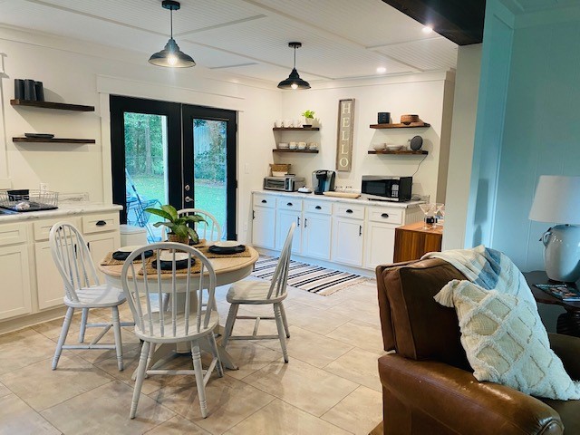 A modern kitchen area features white cabinetry, open shelving, and dark countertops. A round dining table with four chairs occupies the space, while a coffee maker and microwave are visible. Natural light streams through large sliding doors that offer a view of the outdoors.