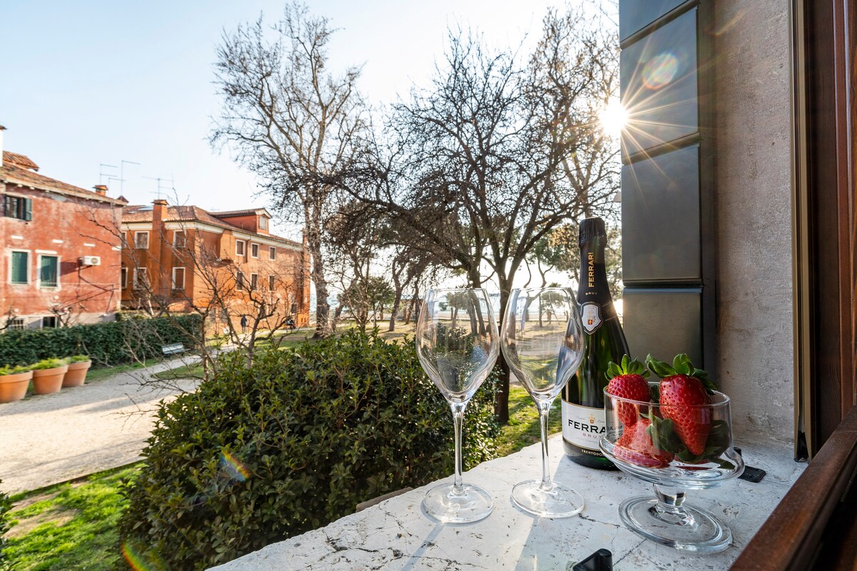 A view from the window showcases a sunlit garden area, framed by trees. A bottle of sparkling wine is displayed alongside two glasses and a bowl of strawberries on the windowsill, creating a serene and inviting scene.