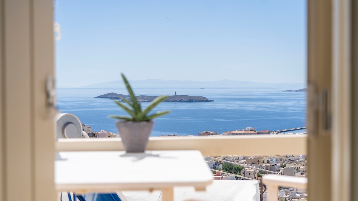 A view of calm blue waters is framed by an open window, showcasing a small island in the distance. A potted plant is placed on a white table, adding a touch of greenery to the serene coastal scene.