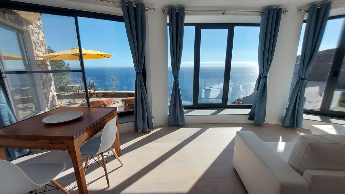 A modern living area is highlighted by large glass windows, offering unobstructed views of the sea. Light filters through sheer curtains, illuminating a wooden dining table surrounded by white chairs. A comfortable sofa is positioned near the window, creating a relaxed environment.