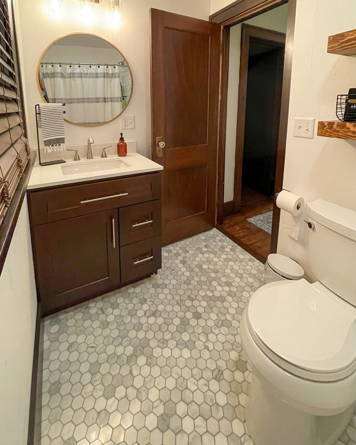 A modern bathroom is presented, featuring a double vanity with two sinks, under-mount lighting, and a round mirror. A stylish hexagonal tile floor complements the neutral color scheme. A shower curtain is visible in the background, and minimal décor is placed on a floating shelf.