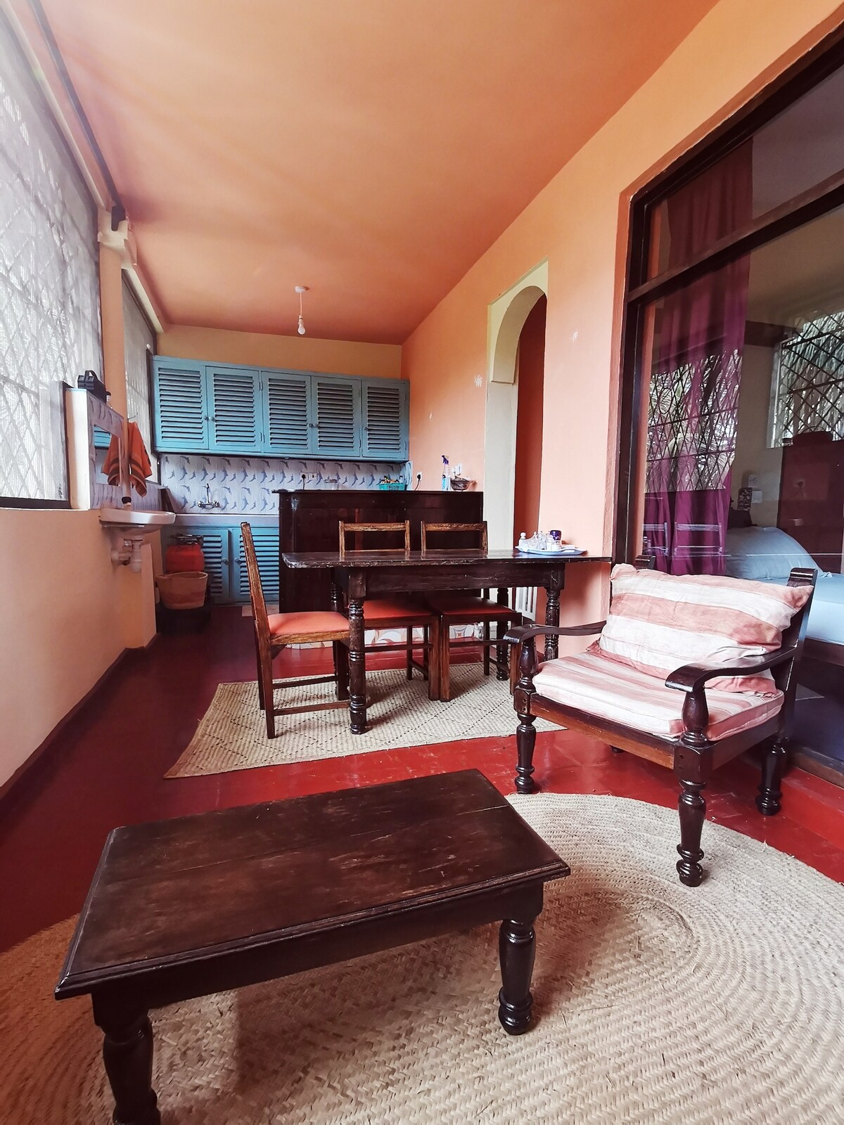 A cozy seating area features a wooden table surrounded by four chairs. A woven rug lies beneath, complementing the hardwood floor. Light enters through large windows adorned with decorative grilles, creating a warm and inviting space.