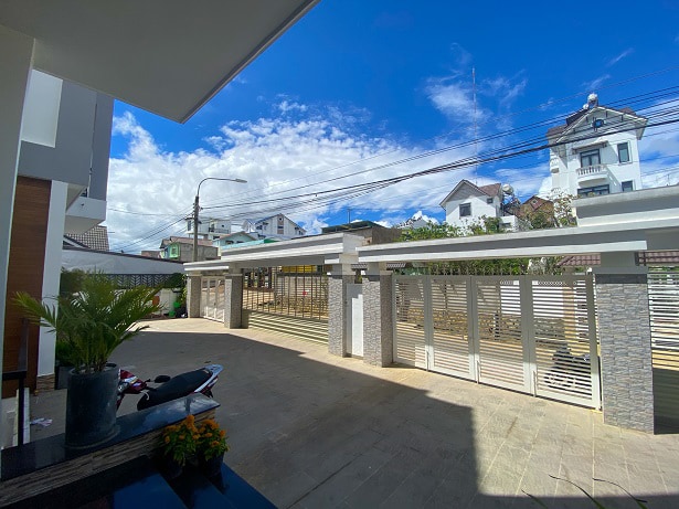 An outdoor space is visible, featuring a stone tiled floor and modern fencing. Lush green plants are arranged in a pot, with a clear blue sky above marked by fluffy white clouds. The area is well-lit and spacious, suitable for gatherings.