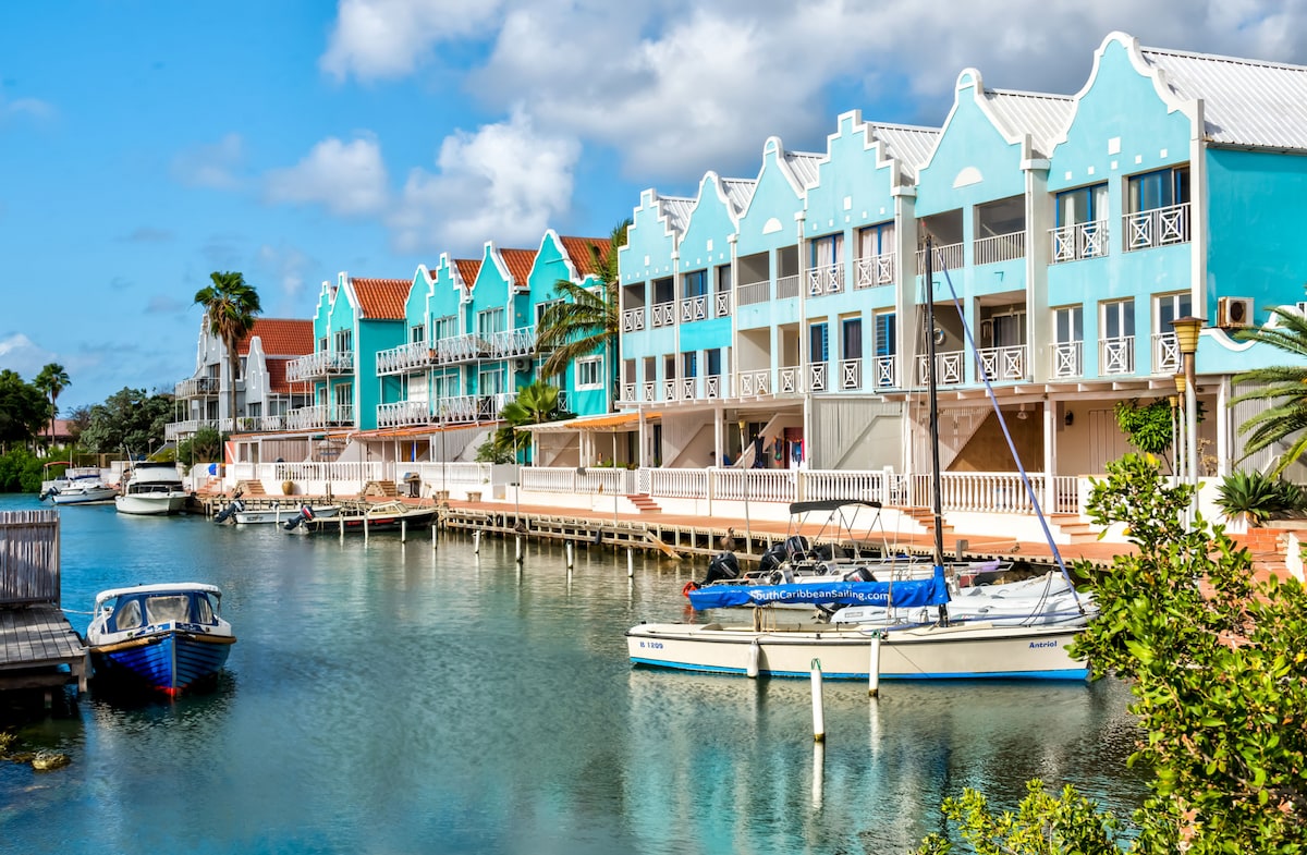 A waterfront view showcases colorful, two-story buildings with distinctive architectural details, lined along a calm canal. Various boats are moored in the foreground, and palm trees add a touch of greenery to the scene, creating a relaxed coastal atmosphere.
