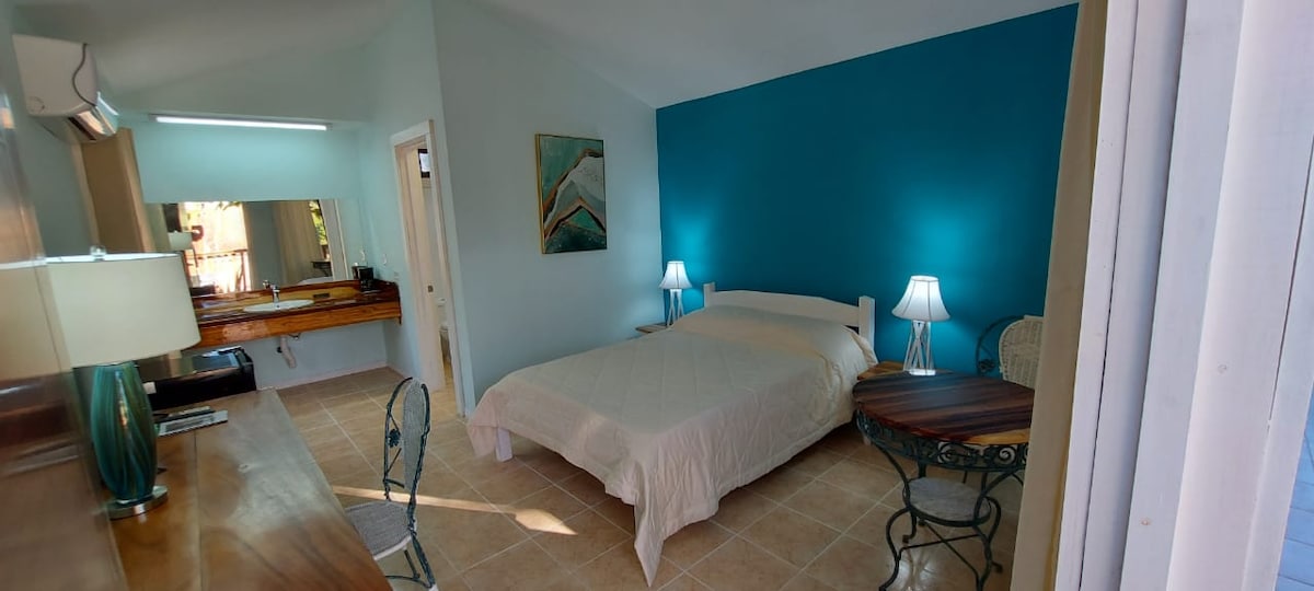 A bright sleeping area features a spacious bed dressed in white linens. Soft turquoise walls provide a tranquil background, while two lamps illuminate either side of the bed. A small dining table and two chairs complement the room's layout, enhancing its functional design.