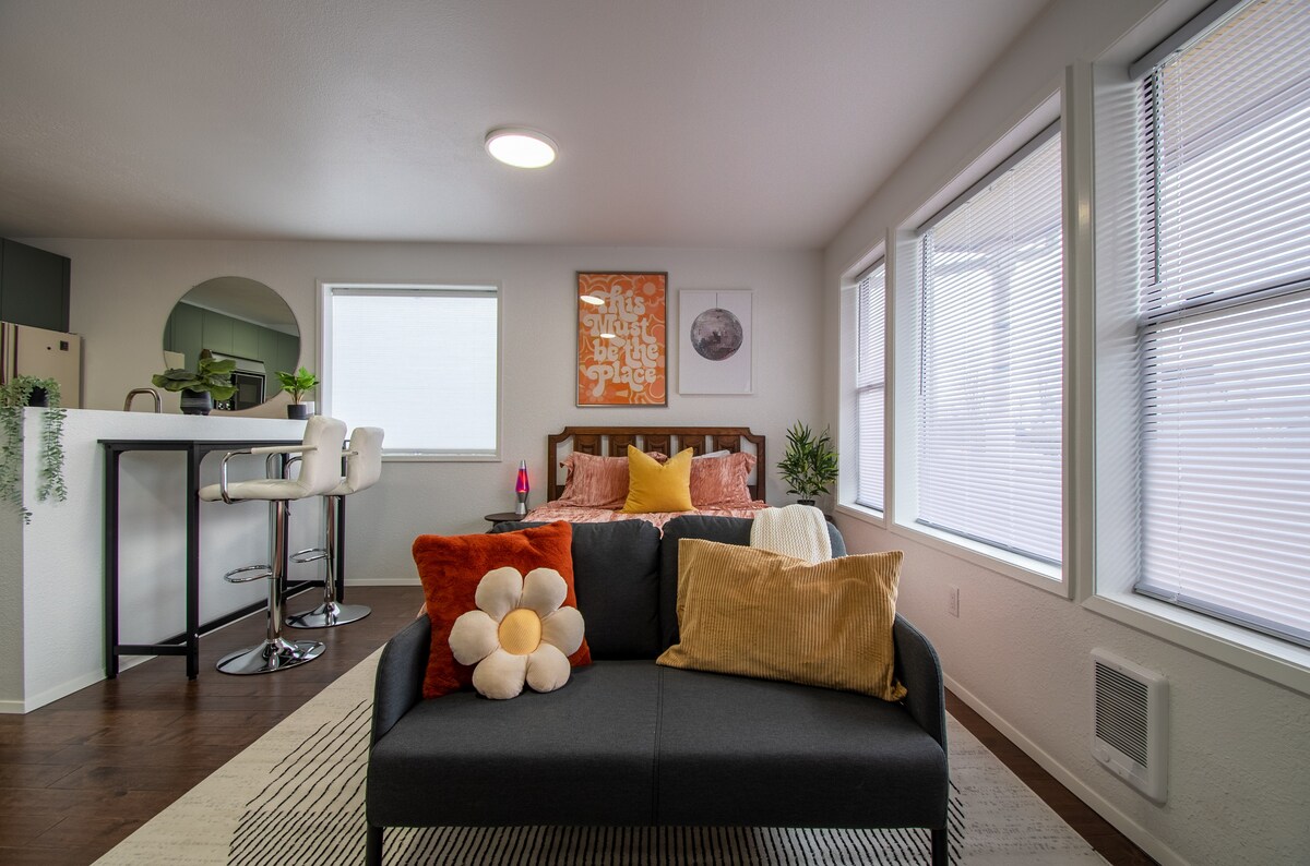 A cozy living space is featured with a dark sofa adorned with colorful cushions, including a flower-shaped pillow. Natural light fills the room through large windows, complemented by light-colored walls and a patterned area rug. A small dining area with bar stools is visible in the background.