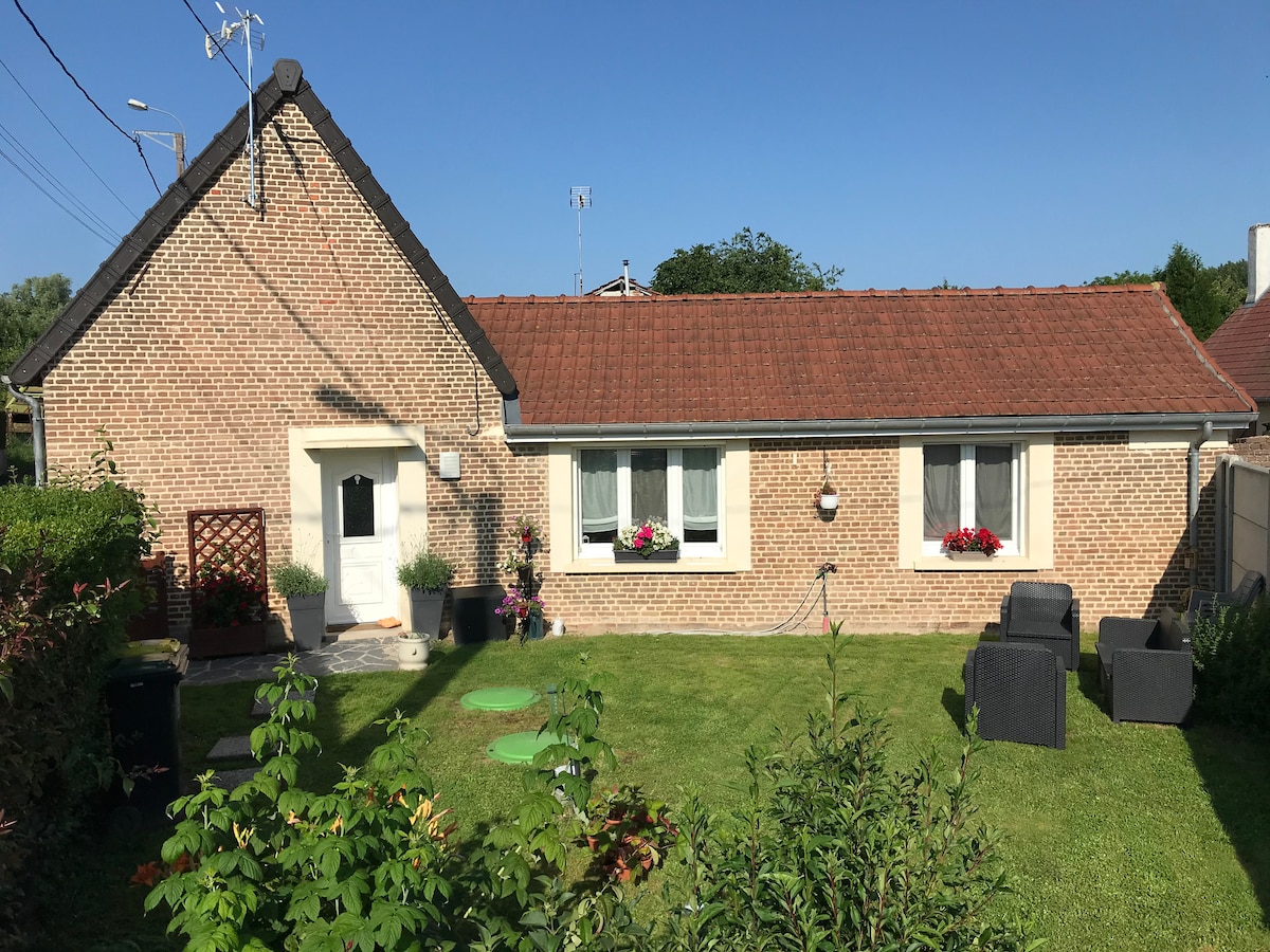 A modern brick house is framed by a well-maintained lawn, featuring flowering plants under the windows. The exterior includes a welcoming entryway and a seating area with dark garden furniture. The clear sky above enhances the inviting setting.
