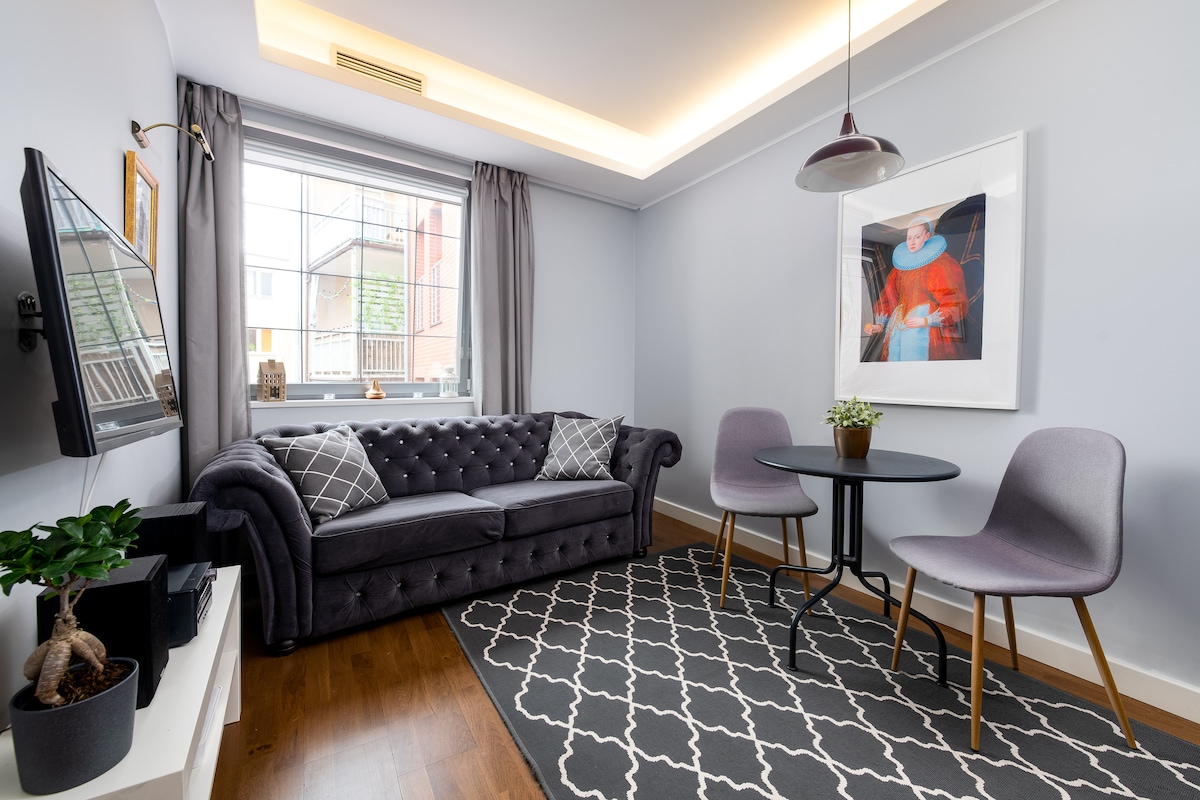 A living area is presented with a plush gray sofa adorned with decorative cushions. A small round table with two chairs is positioned nearby. A television is mounted on the wall, and light enters through a window draped in gray curtains. A potted plant adds a touch of greenery.