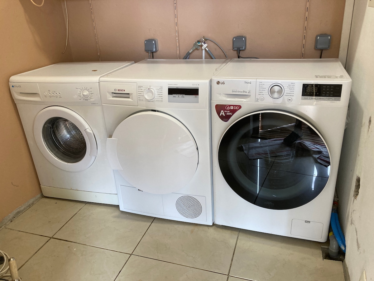 A laundry area is equipped with three white appliances: a washing machine, a condenser dryer, and an additional washing machine. The appliances are neatly arranged on a tiled floor, with minimal wall space visible behind them.