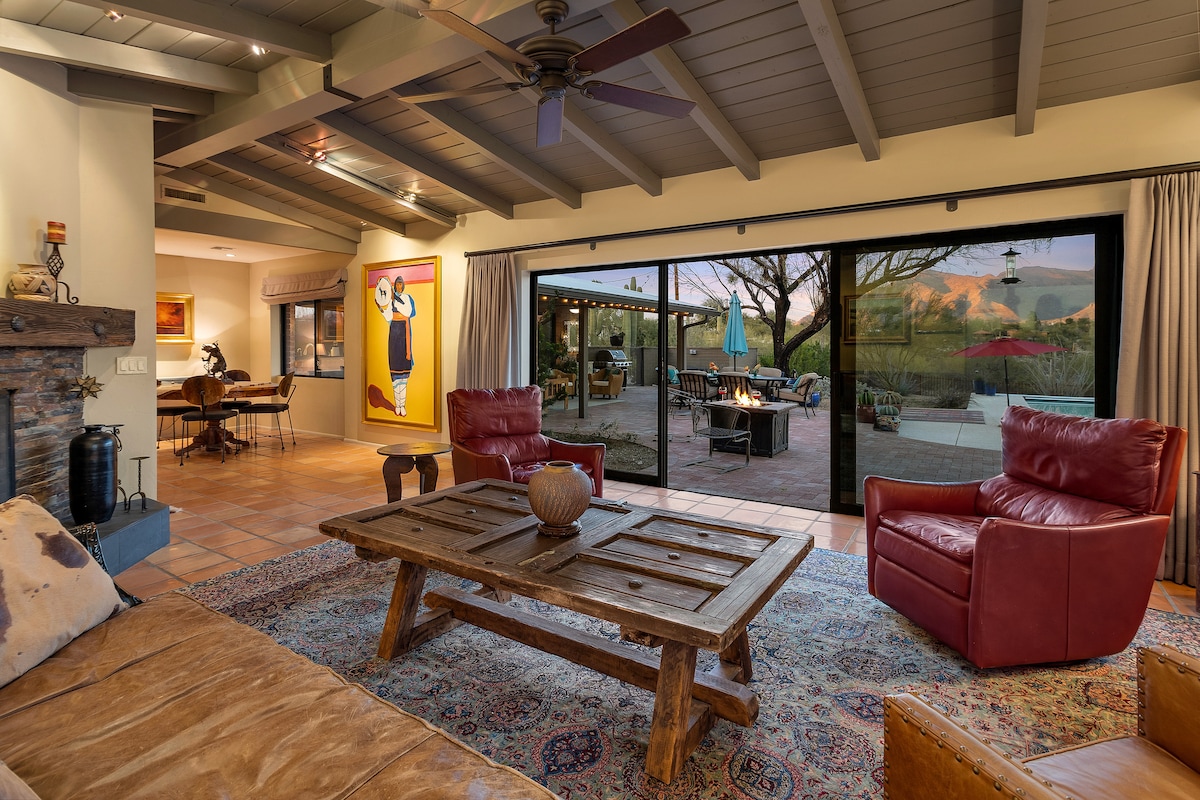 A spacious living room features a rustic wooden coffee table at its center, surrounded by comfortable seating. Large glass doors open to a picturesque outdoor space, and a colorful wall art piece adds charm to the atmosphere. Natural light illuminates the room.