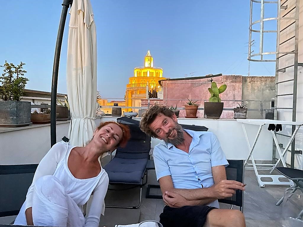 The terrace features comfortable loungers and a large umbrella providing shade. In the background, a notable building is illuminated, adding character to the evening sky. Potted plants are arranged atop the terrace railing, enhancing the outdoor atmosphere.