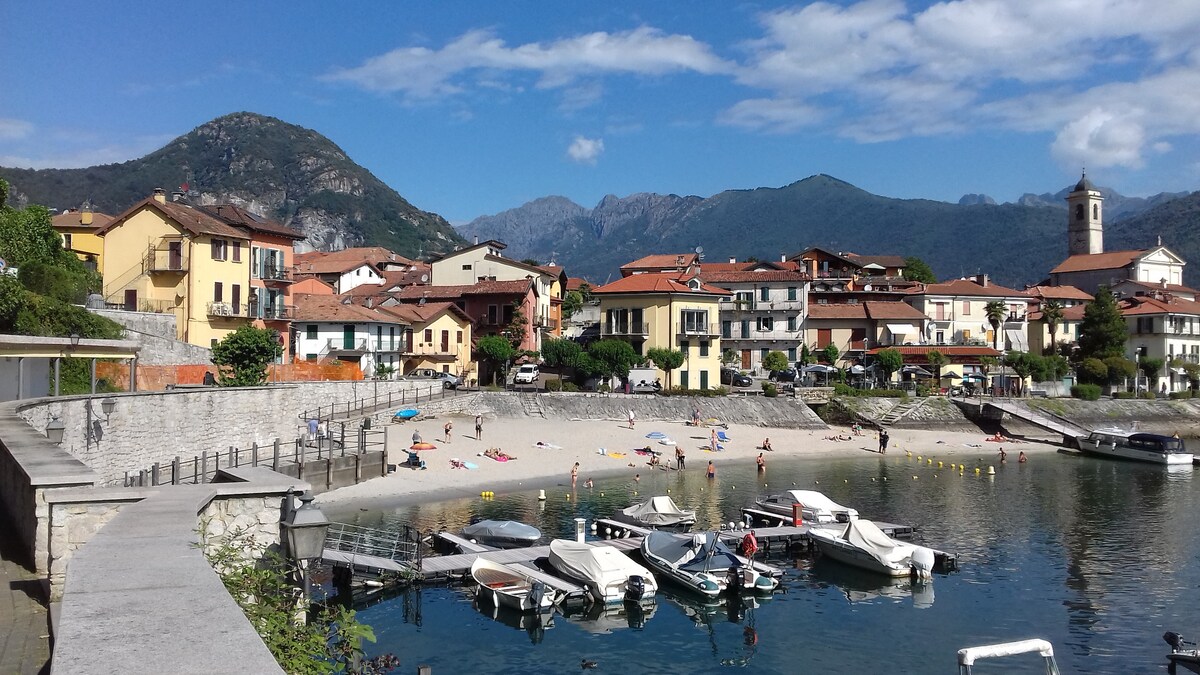 A serene lakeside view showcases a beach with sunbathers and colorful umbrellas. Numerous boats are docked in the clear water, while charming buildings with red roofs line the shore. The backdrop features lush mountains under a clear sky.
