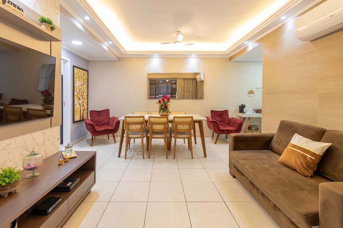 A spacious living and dining area is shown, featuring a dining table with six chairs, a comfortable brown sofa, and two red armchairs. A smart TV is mounted on the wall, with soft lighting highlighting the decor. A ceiling fan enhances airflow within the space.