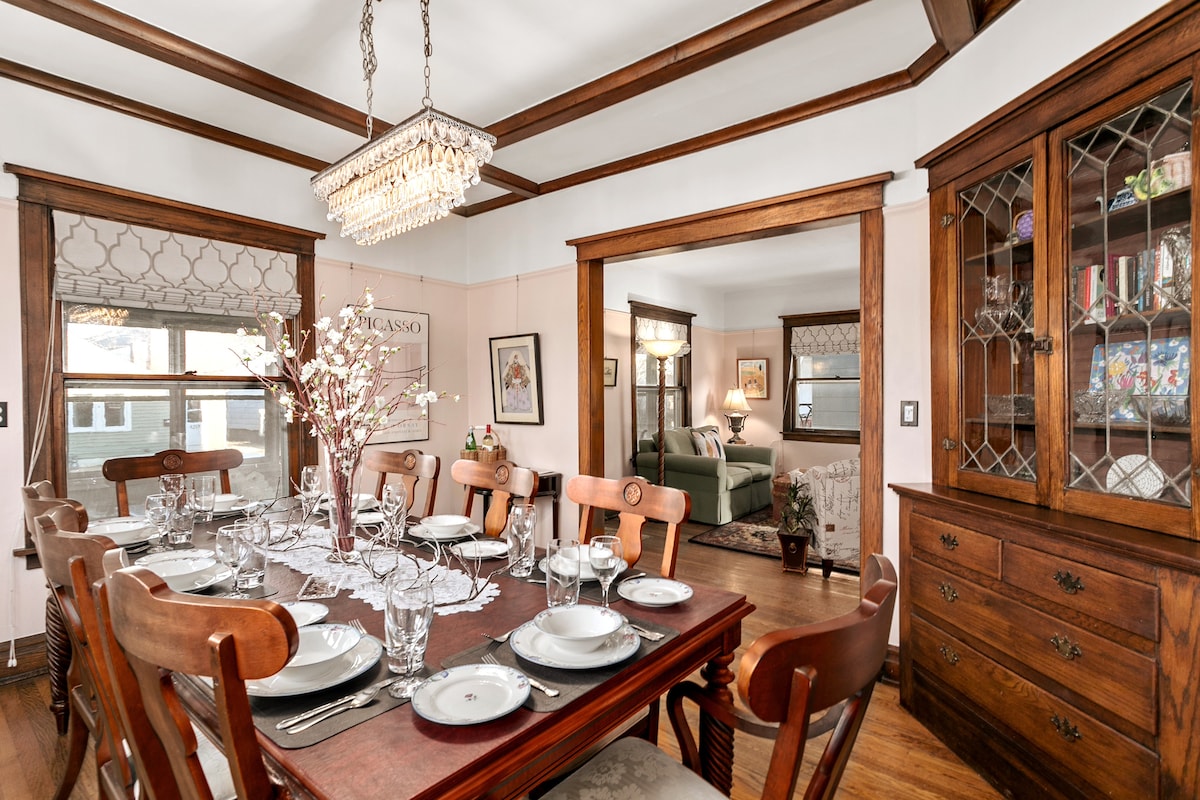 A spacious dining area showcases a large wooden table set for a meal, featuring elegant dinnerware and glassware. Classic wooden beams frame the room, complemented by a crystal chandelier above. A cabinet houses decorative items and provides a warm, inviting atmosphere.