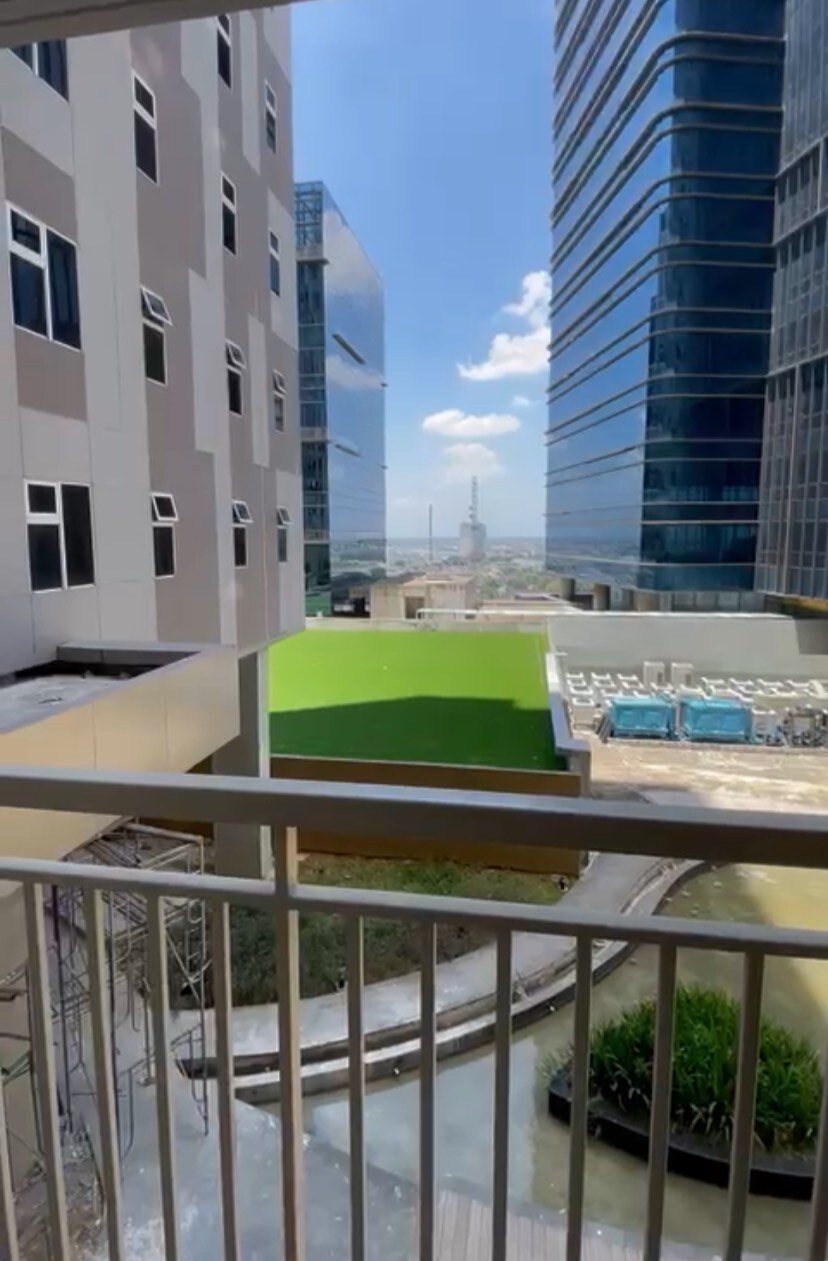 A balcony view showcases a green outdoor space and swimming pool below, with tall buildings visible in the background. The sky appears bright and clear, allowing for ample natural light in the surroundings.