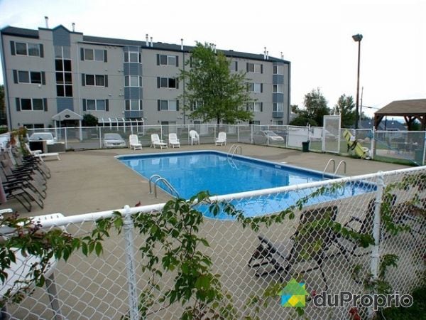 An outdoor swimming pool is surrounded by a fenced area, featuring a concrete deck with multiple lounge chairs. The backdrop includes a three-story building, and greenery is visible nearby, enhancing the setting.