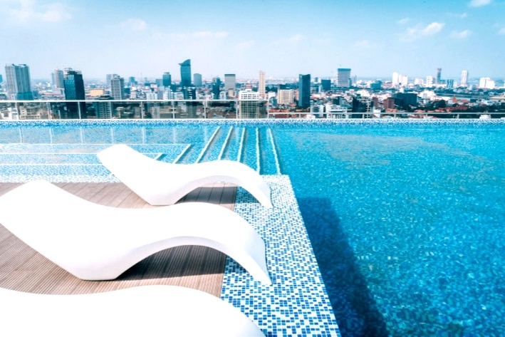 An infinity rooftop pool is depicted, featuring sleek white lounge chairs positioned along the edge. The shimmering water reflects the bright sky, with a panoramic view of the Phnom Penh skyline visible in the background.