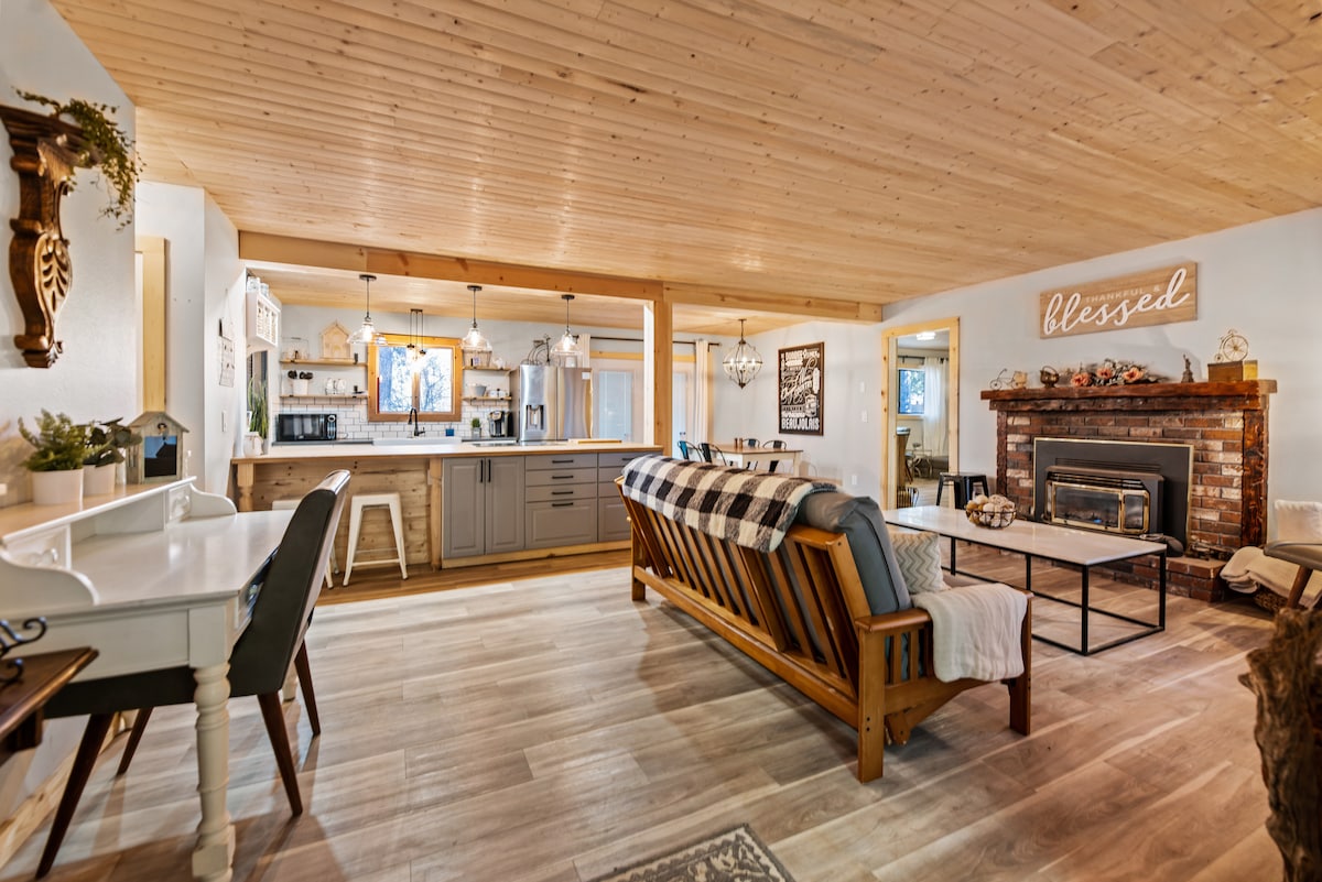 A spacious living area featuring wood paneling and a high ceiling. A comfortable sofa with throw blankets faces a brick fireplace. Adjacent, a kitchen with modern appliances and a dining area can be seen. Subtle decor elements add a welcoming touch to the space.