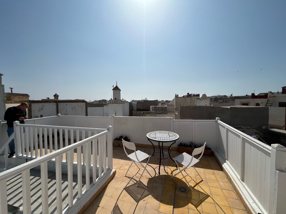 A rooftop terrace is shown under a bright blue sky, featuring a round table surrounded by two white chairs. A detailed view of the surrounding architecture and rooftops of Essaouira can be seen in the background, providing a serene outdoor setting.