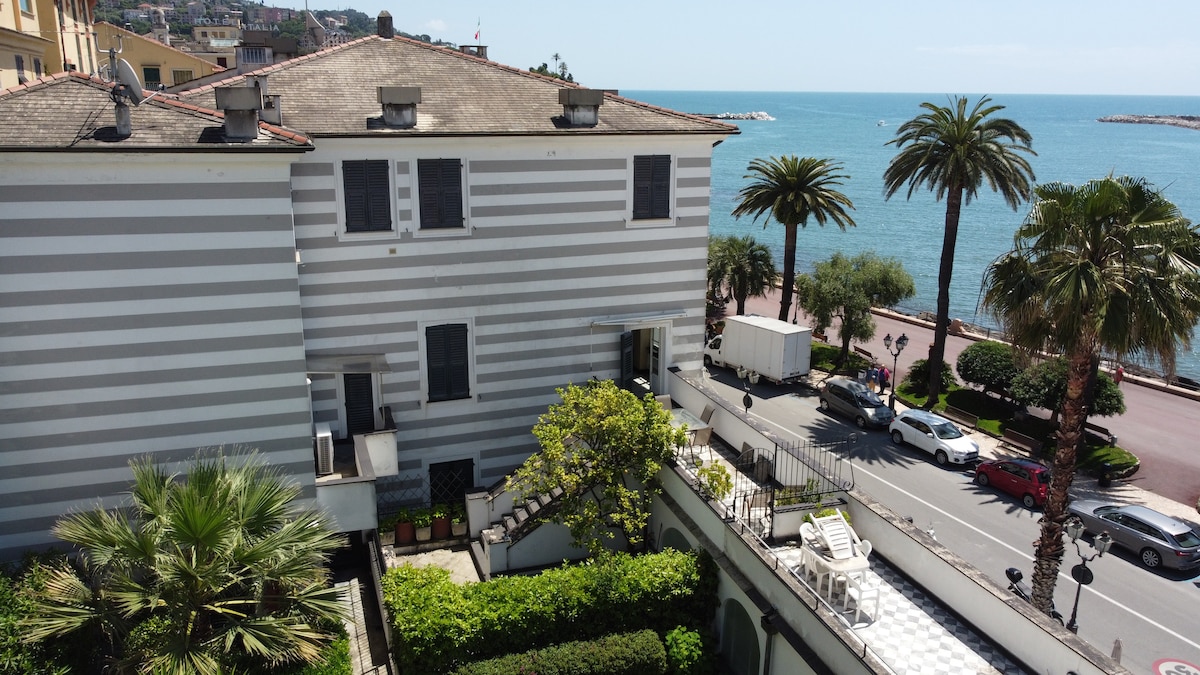 The image captures a classic building with striped gray and white exterior, located adjacent to the coastline. Palm trees and lush greenery are visible in the foreground, while the serene blue sea is seen in the background, suggesting a tranquil atmosphere near the waterfront.
