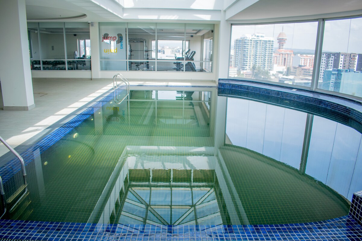 A modern heated swimming pool is situated indoors with large windows that provide a panoramic view of the city skyline. Natural light reflects off the water's surface, creating a serene ambiance. The surrounding area features fitness equipment visible in the background.