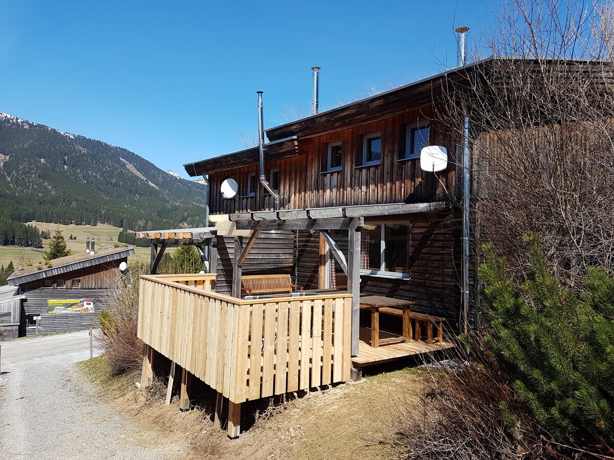 A wooden chalet is set against a clear blue sky, featuring a spacious terrace with wooden railings. The structure is surrounded by greenery and mountain landscapes, showcasing its natural setting. Outdoor seating is visible on the terrace, inviting relaxation in a serene environment.