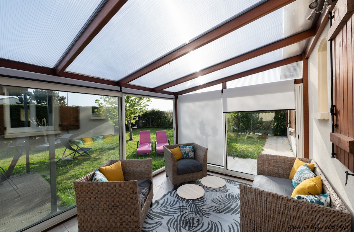 A sunlit veranda features large glass panels, offering views of the garden. Two comfortable wicker chairs with colorful cushions are arranged around a round rug. Outside, lounge chairs and a table can be seen, creating a welcoming outdoor space.