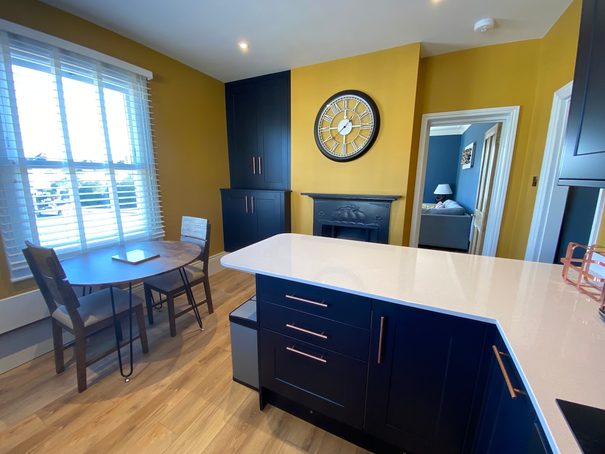 A modern kitchen area is highlighted by a round dining table with four chairs. A large window with white blinds allows natural light to fill the space. Dark cabinetry contrasts with a bright countertop. A decorative clock is mounted on the wall.