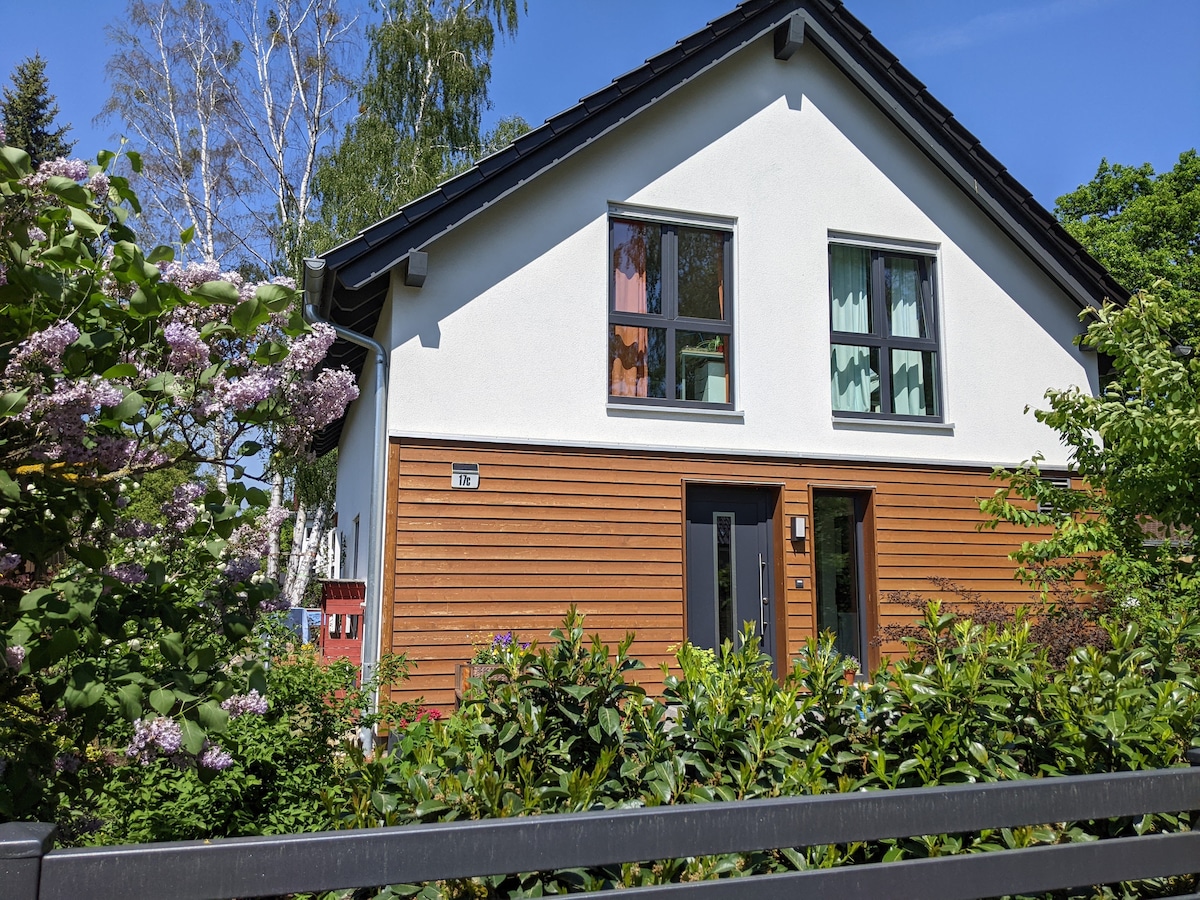 A modern, two-story house is surrounded by greenery, featuring a combination of white and natural wood siding. Large windows allow natural light to fill the interior. Colorful curtains are visible through the windows, enhancing the exterior with a sense of liveliness.
