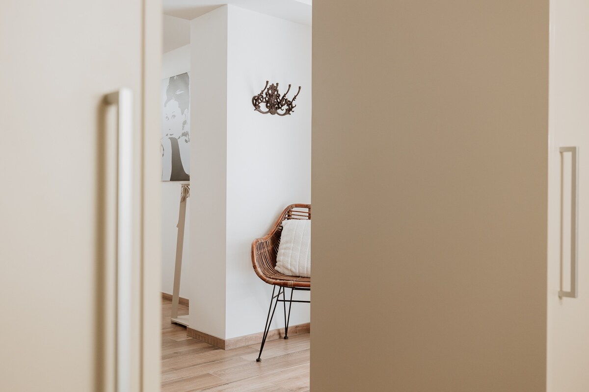 The entryway opens into a light-filled space featuring a rattan chair positioned against a neutral wall. Simple decor elements create a sense of warmth. Visible flooring is crafted from light-colored wood, enhancing the overall clean and contemporary feel of the apartment.