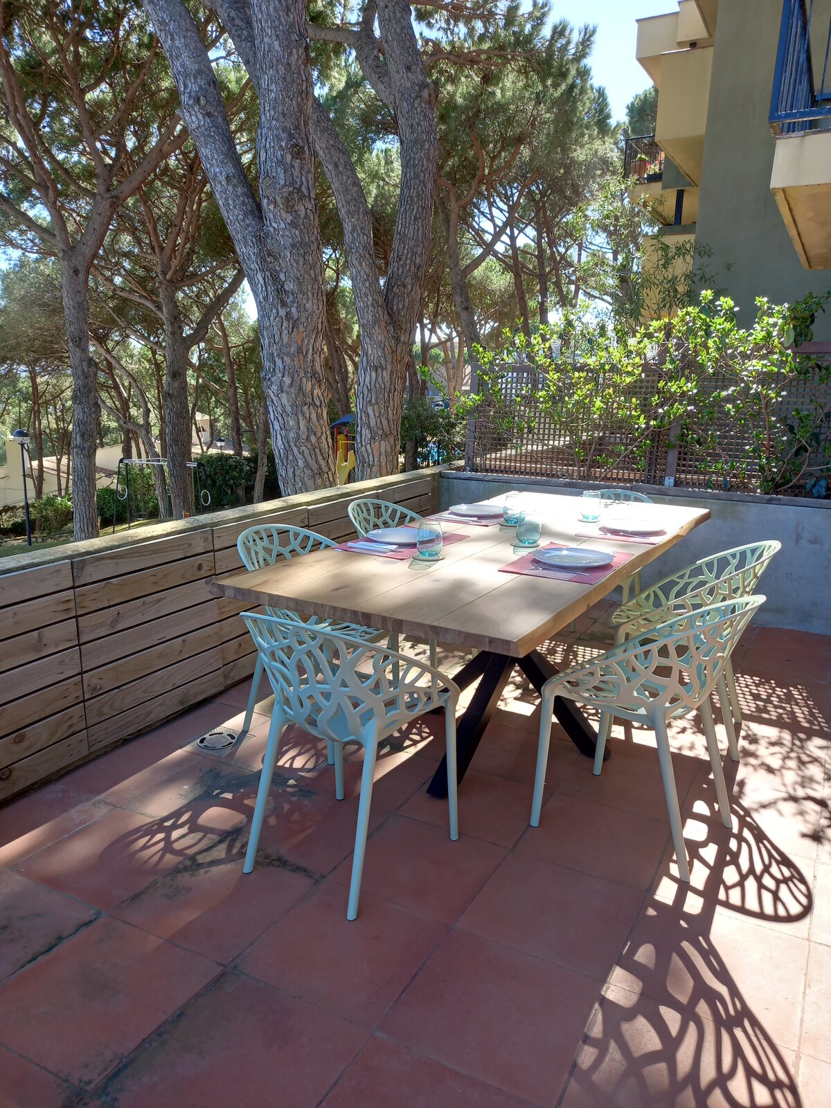An outdoor dining area features a large wooden table surrounded by six light-colored chairs. Tall trees provide shade in the background, creating a serene environment. The table is set with plates and utensils, indicating a space prepared for meals shared with family and friends.