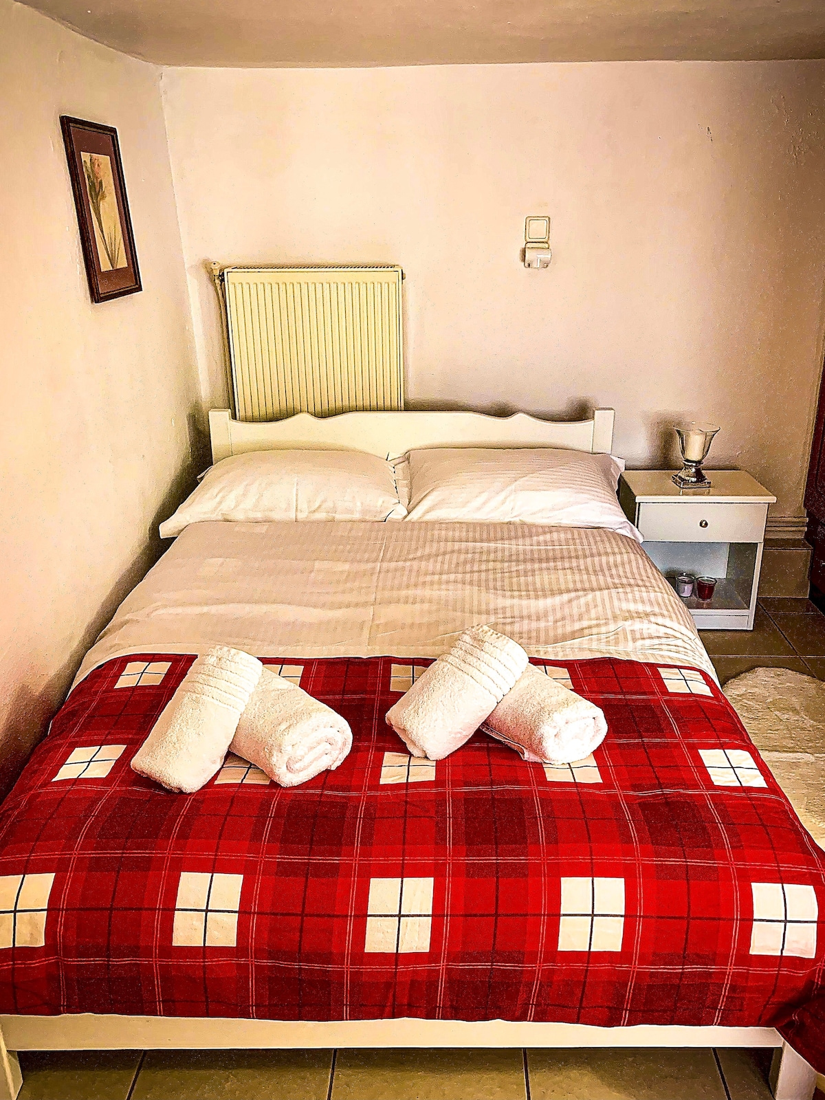 A compact bedroom features a double bed dressed with crisp linens and a vibrant red plaid blanket. Fresh towels are neatly placed at the foot of the bed. A small nightstand stands beside the bed with a subtle decorative item and soft lighting from a nearby wall fixture.