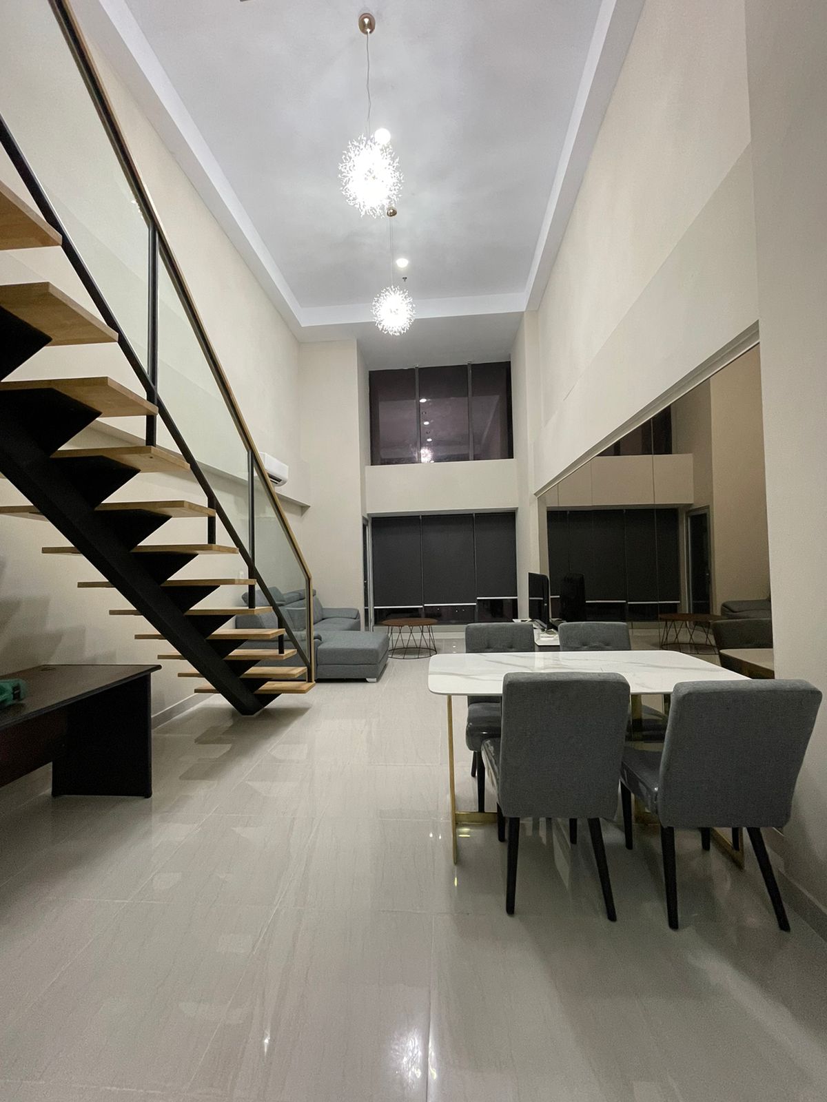 A spacious living area is highlighted by high ceilings and modern lighting fixtures. A staircase ascends to the upper level. A dining table with four chairs is positioned beside a large window, where natural light enhances the neutral-toned decor.