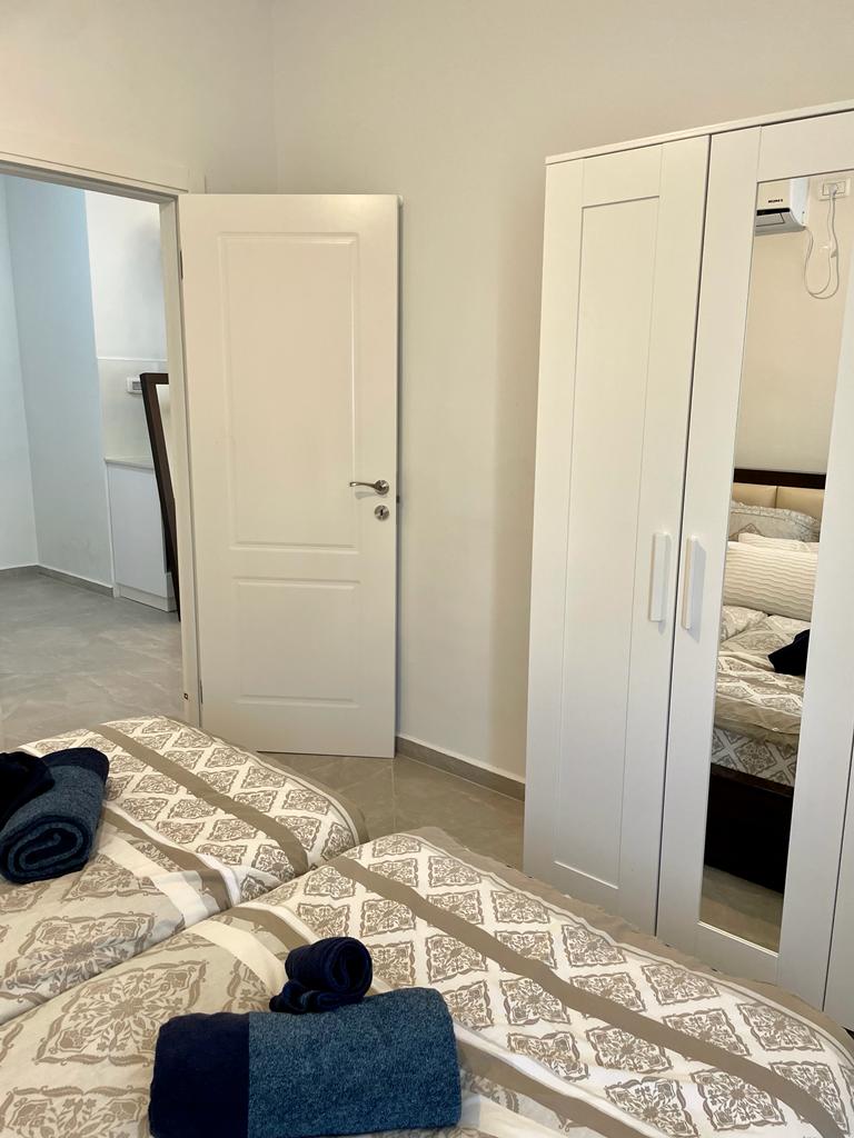 A light-filled bedroom features two twin beds with decorative bedding. A mirror-fronted wardrobe stands against the wall, reflecting the room's neutral tones. Fresh towels are neatly placed on each bed, contributing to an organized and restful environment.