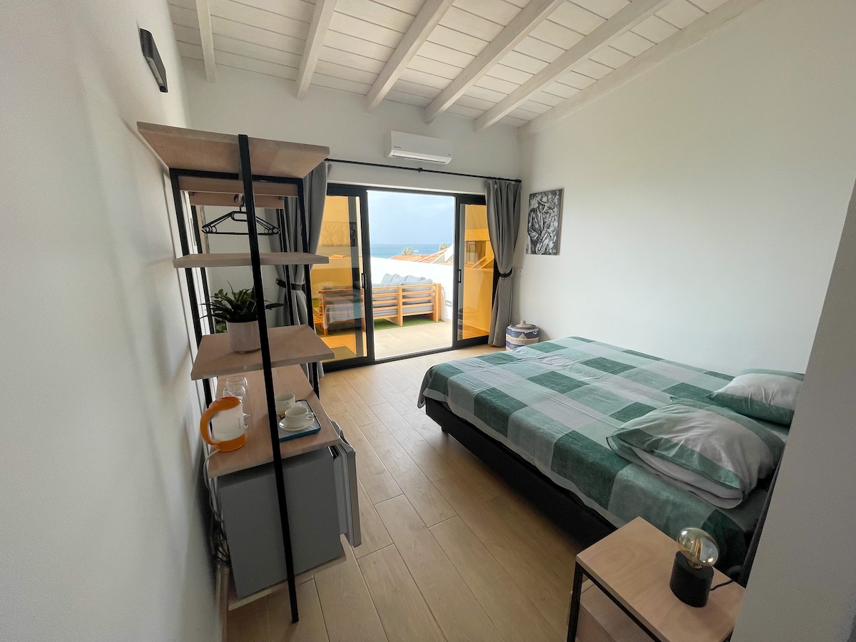 A modern bedroom features a spacious bed with green and gray bedding. A shelving unit holds a small refrigerator and kettle. Large glass doors lead to a shared outdoor terrace, allowing natural light to fill the room.