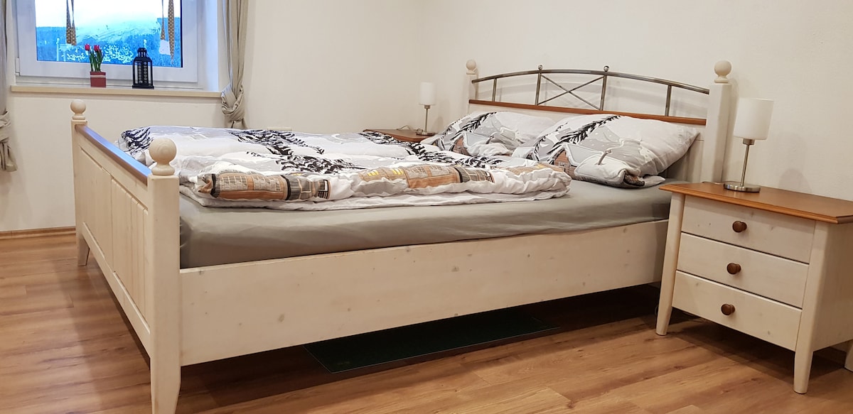 A spacious bed with a light-colored wooden frame is positioned in a well-lit room. Soft bedding in neutral tones adds comfort. A bedside table with a lamp is placed on one side, complementing the serene atmosphere, while a window allows natural light to enter.