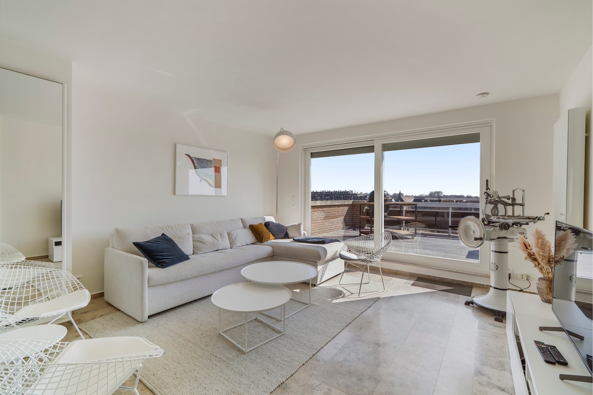 A spacious living area is highlighted by a large window that offers natural light and views of the outdoor terrace. A comfortable sofa, paired with modern accent chairs, surrounds a coffee table, creating an inviting space for relaxation. The overall decor is minimalist and stylish.