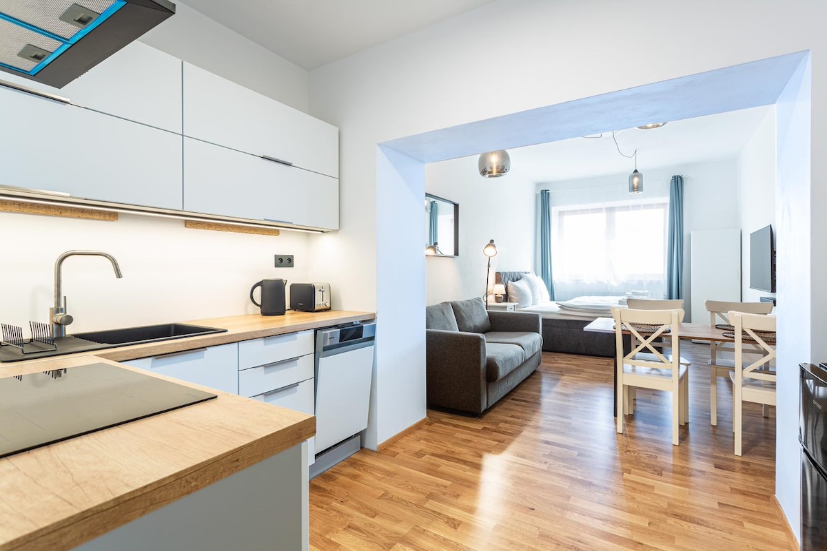 A modern kitchen space is equipped with sleek cabinets, a stove, and a dishwasher, extending into a living area featuring a comfortable sofa and a dining table with four chairs. Large windows allow natural light to fill the room.