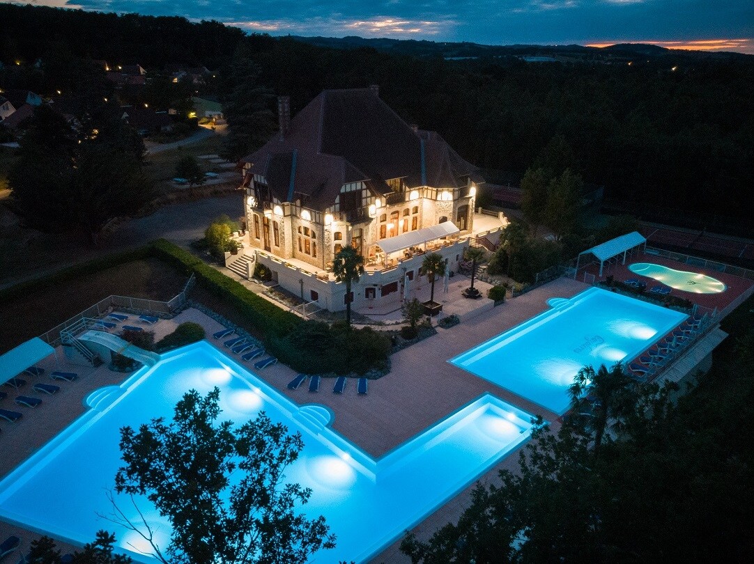 An aerial view captures a well-lit building surrounded by two illuminated swimming pools during twilight. Palm trees provide greenery, and lounge chairs line the poolside, enhancing the inviting ambiance of the vacation park.