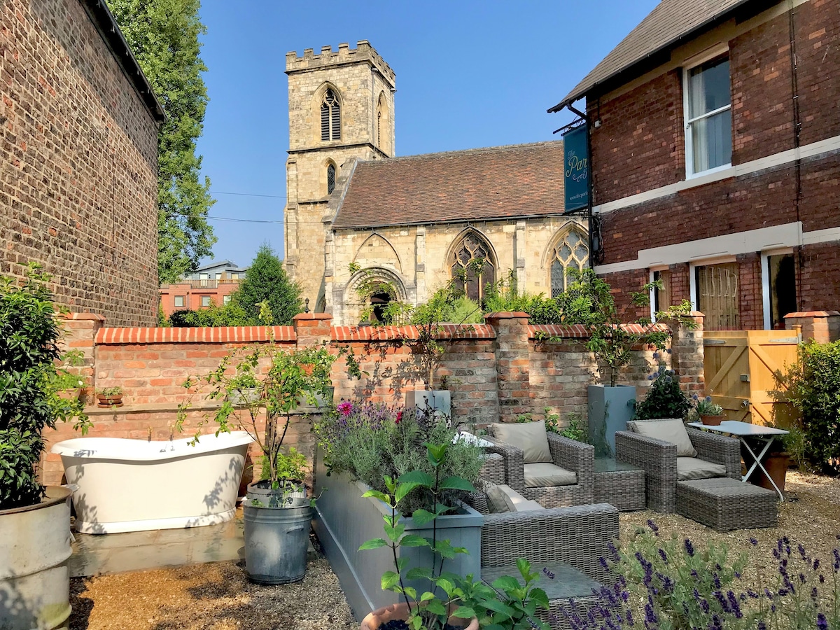 A private garden features a relaxing outdoor bath, surrounded by a variety of potted plants and low-maintenance shrubs. Comfortable seating areas with woven furniture are positioned on gravel, providing a tranquil space. A historic church tower and clear blue sky are visible in the background.