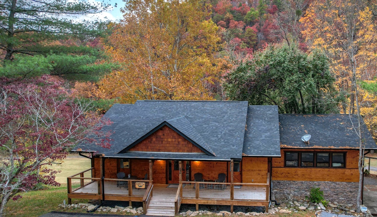 The exterior of the lodge displays a blend of natural wood and stone, surrounded by vibrant fall foliage. A welcoming porch features seating, and the surrounding landscape includes trees and grassy areas, creating a serene environment. Mountains are visible in the background.