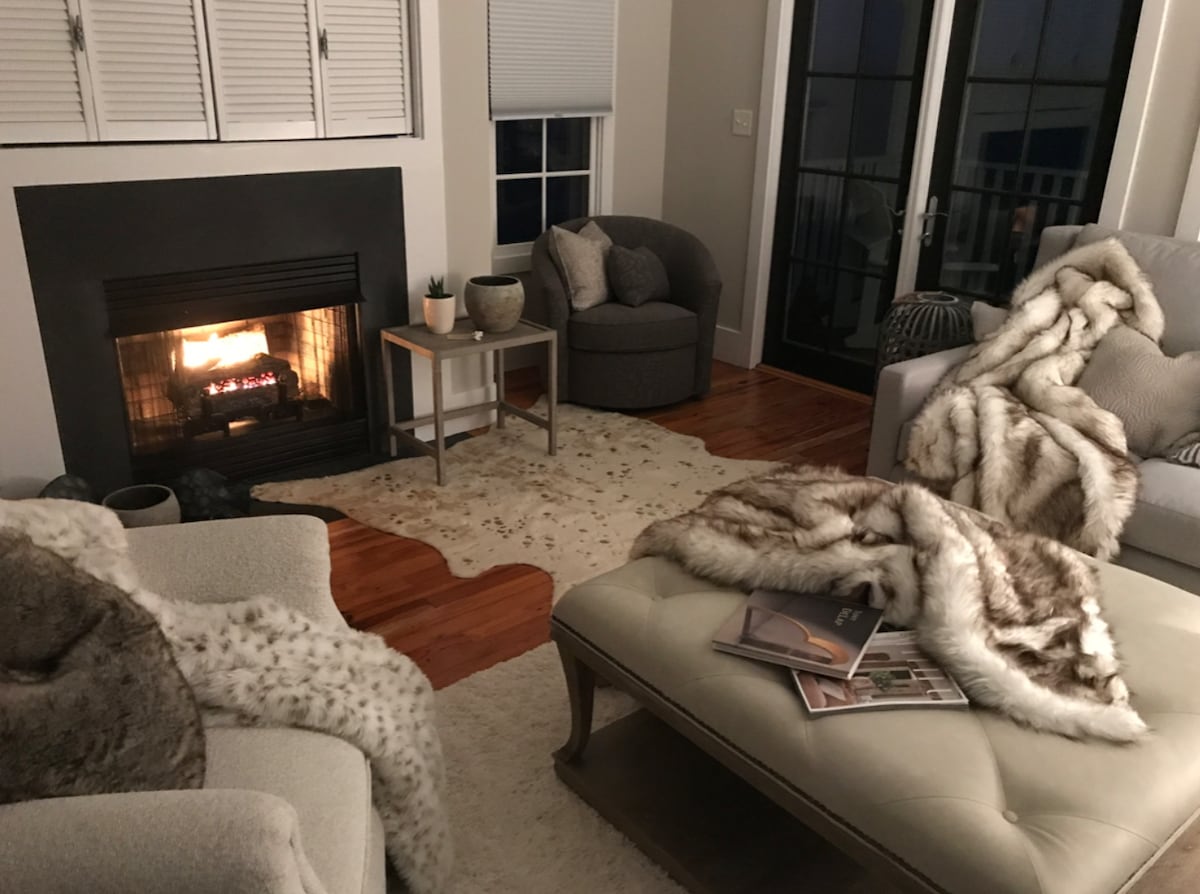 A cozy living area features a comfortable fireplace, surrounded by soft seating options and a light area rug. Textured throws are casually placed on the chairs and ottoman, creating an inviting ambiance. Large glass doors provide access to an outdoor balcony.
