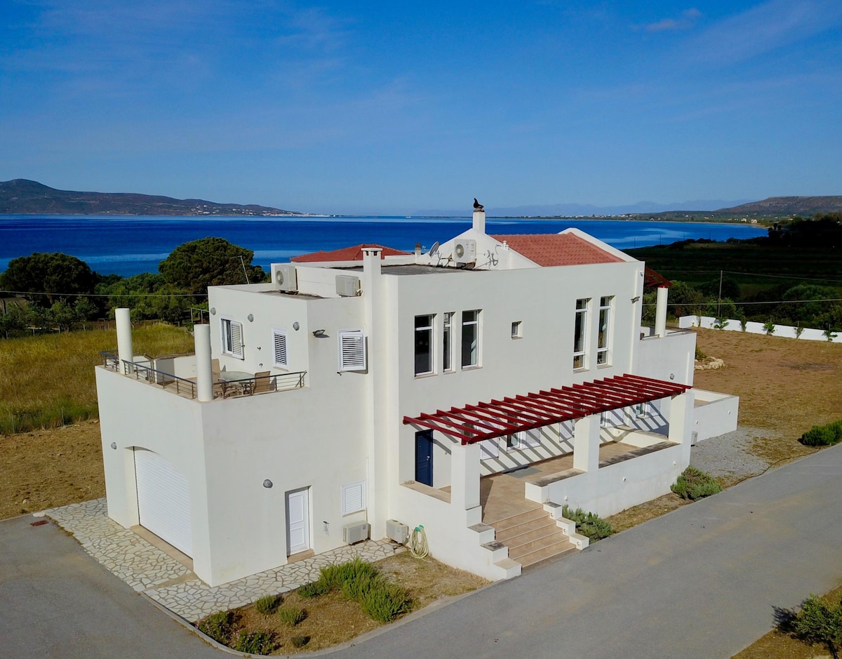 A light-colored, modern home is positioned along a serene coastal landscape. The building features multiple terraces and large windows that provide clear views of the sea. Surrounding greenery and a paved pathway lead to the entrance, enhancing the inviting exterior.