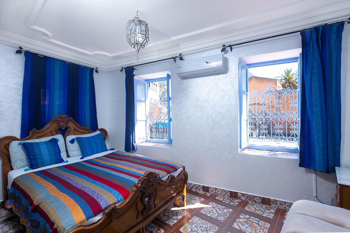 A cozy bedroom is illuminated by natural light from two windows adorned with bright blue curtains. A spacious bed with a colorful striped blanket is positioned against a wall, complemented by decorative pillows. Air conditioning and intricate tile flooring are also present.