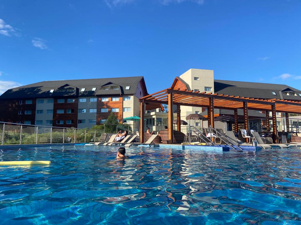 A large outdoor pool is surrounded by sun loungers, with a building featuring a mix of natural wood and concrete elements in the background. Clear blue water reflects the sky, and a person is seen swimming, emphasizing a welcoming and relaxed environment.