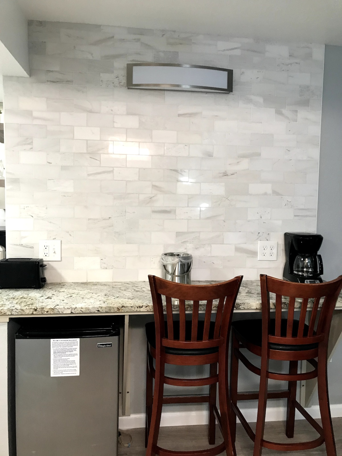 A kitchen bar area is depicted, featuring two wooden stools positioned beside a granite countertop. The backsplash is designed with white marble tiles. A compact refrigerator and a coffee maker are visible on the countertop, providing functional amenities.
